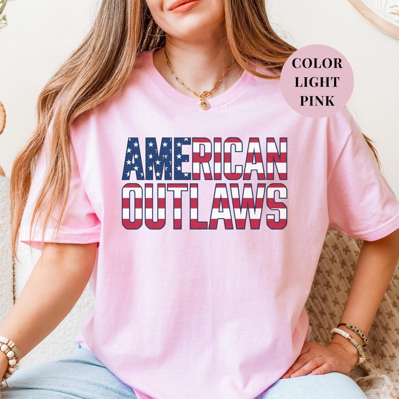 Pink t-shirt featuring a distressed retro design for "AMERICAN OUTLAWS". The bold collegiate block lettering is filled with a patriotic US national flag pattern of stars and stripes. High-quality vintage-style soccer apparel for passionate United States football supporters and tournament fans.