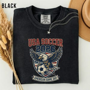 May include: Black t-shirt with a graphic featuring an eagle holding a soccer ball, the text "USA SOCCER 2026," and "AMERICAN GAME DAY." A silver star necklace and a brown hat are visible.