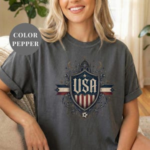 A woman wearing a gray t-shirt with a graphic design featuring the text "USA" and a shield-like emblem with stars and stripes.