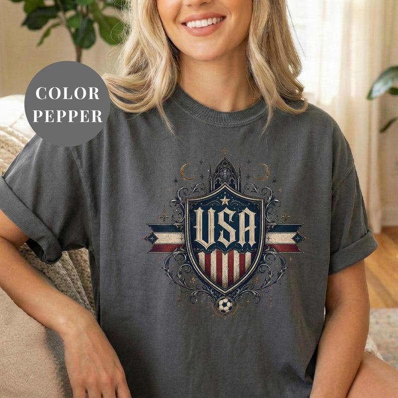 A woman wearing a gray t-shirt with a graphic design featuring the text "USA" and a shield-like emblem with stars and stripes.
