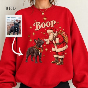Puede incluir: Una sudadera roja de cuello redondo con un gráfico de Santa Claus y un bulldog francés negro con una corona de cascabeles. El texto BOOP está impreso encima, rodeado de estrellas doradas. Una pequeña foto insertada muestra al perro.