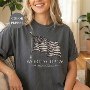 Pepper Comfort Colors 1717 t-shirt featuring a vintage hand-drawn sketch of a waving American flag. Includes "WORLD CUP '26" and "United States" text, with a subtle soccer ball detail on the flagpole. High-quality patriotic soccer apparel for the 2026 world tournament fans.