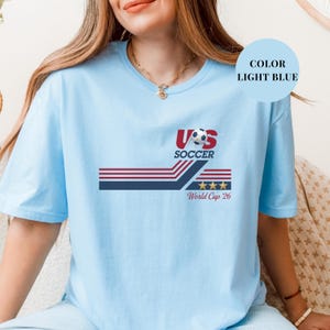 A woman wearing a light blue t-shirt with a graphic design featuring a soccer ball and the text "US SOCCER World Cup 26".