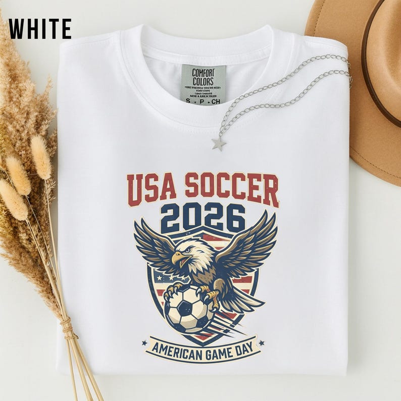 May include: White t-shirt with the text "USA SOCCER 2026" in red and blue. The shirt features an eagle graphic holding a soccer ball, with the words "AMERICAN GAME DAY" below. A silver necklace with a star pendant is on the shirt.
