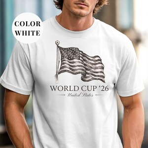 White Comfort Colors 1717 t-shirt featuring a vintage hand-drawn sketch of a waving American flag. Includes "WORLD CUP '26" and "United States" text, with a subtle soccer ball detail on the flagpole. High-quality patriotic soccer apparel for the 2026 world tournament fans.