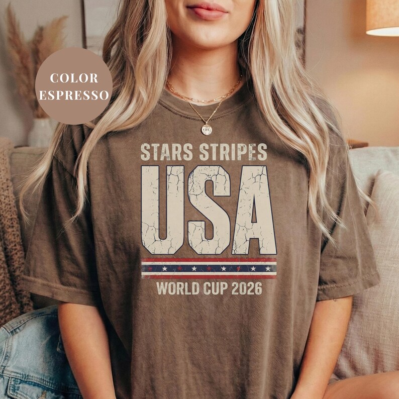 Espresso brown t-shirt featuring a distressed vintage "STARS STRIPES USA WORLD CUP 2026" graphic. The design includes weathered block lettering for "USA" and a patriotic red, white, and blue banner with stars. High-quality sports apparel for United States soccer fans and world tournament supporters.