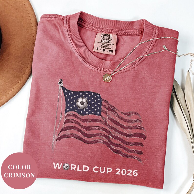 Crimson Comfort Colors 1717 t-shirt featuring a vintage-style waving American flag. The design includes a soccer ball integrated into the stars and "WORLD CUP 2026" bold text. High-quality patriotic sports apparel for United States soccer fans and tournament supporters.
