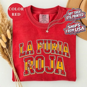 May include: A red t-shirt with the text "LA FURIA ROJA" in yellow and red lettering. The shirt has a crew neck and short sleeves. A small silver necklace with a star pendant is visible. The shirt is labeled "Comfort Colors".