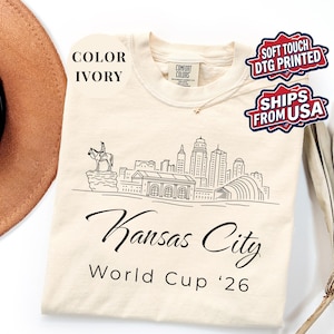 Ivory Comfort Colors T-shirt featuring a minimalist black line art Kansas City skyline design, elegant Kansas City script lettering, and World Cup ’26 text, styled as a soccer travel souvenir shirt for Kansas City fans, football lovers, and 2026 World Cup city apparel.