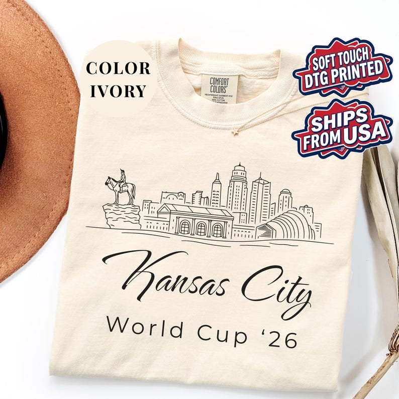Ivory Comfort Colors T-shirt featuring a minimalist black line art Kansas City skyline design, elegant Kansas City script lettering, and World Cup ’26 text, styled as a soccer travel souvenir shirt for Kansas City fans, football lovers, and 2026 World Cup city apparel.