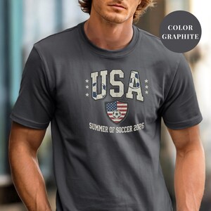 Graphite Comfort Colors 1717 t-shirt with a vintage "USA Summer of Soccer 2026" graphic. Features bold varsity "USA" lettering with soccer ball patterns and a distressed American flag soccer shield. Patriotic apparel for World Cup 2026 fans.