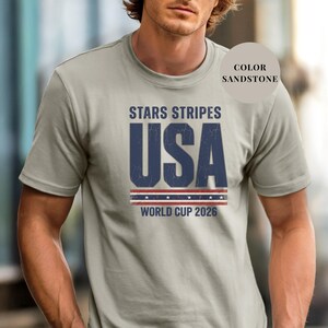 Sand t-shirt featuring a distressed vintage "STARS STRIPES USA WORLD CUP 2026" graphic. The design includes weathered block lettering for "USA" and a patriotic red, white, and blue banner with stars. High-quality sports apparel for United States soccer fans and world tournament supporters.