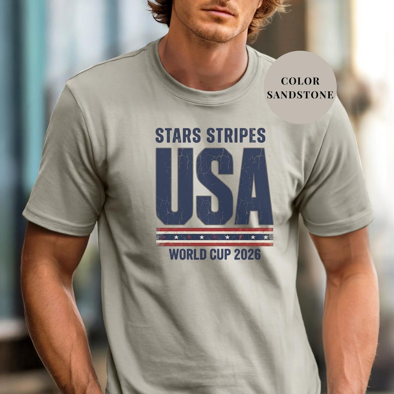 Sand t-shirt featuring a distressed vintage "STARS STRIPES USA WORLD CUP 2026" graphic. The design includes weathered block lettering for "USA" and a patriotic red, white, and blue banner with stars. High-quality sports apparel for United States soccer fans and world tournament supporters.