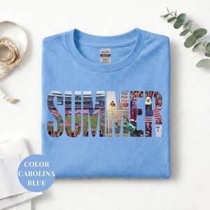 Blue Gildan t-shirt featuring a "SUMMER" photo collage graphic. The bold block letters are filled with USA soccer scenes, including a packed stadium, American flags, cheering fans, and a diving goalie. Patriotic sports apparel for World Cup 2026 supporters.