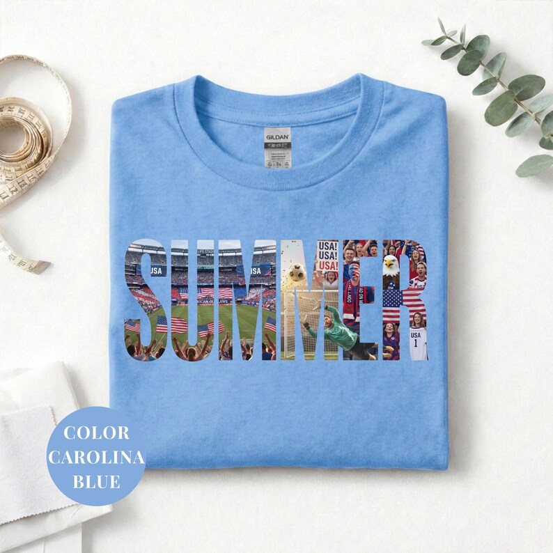 Blue Gildan t-shirt featuring a "SUMMER" photo collage graphic. The bold block letters are filled with USA soccer scenes, including a packed stadium, American flags, cheering fans, and a diving goalie. Patriotic sports apparel for World Cup 2026 supporters.