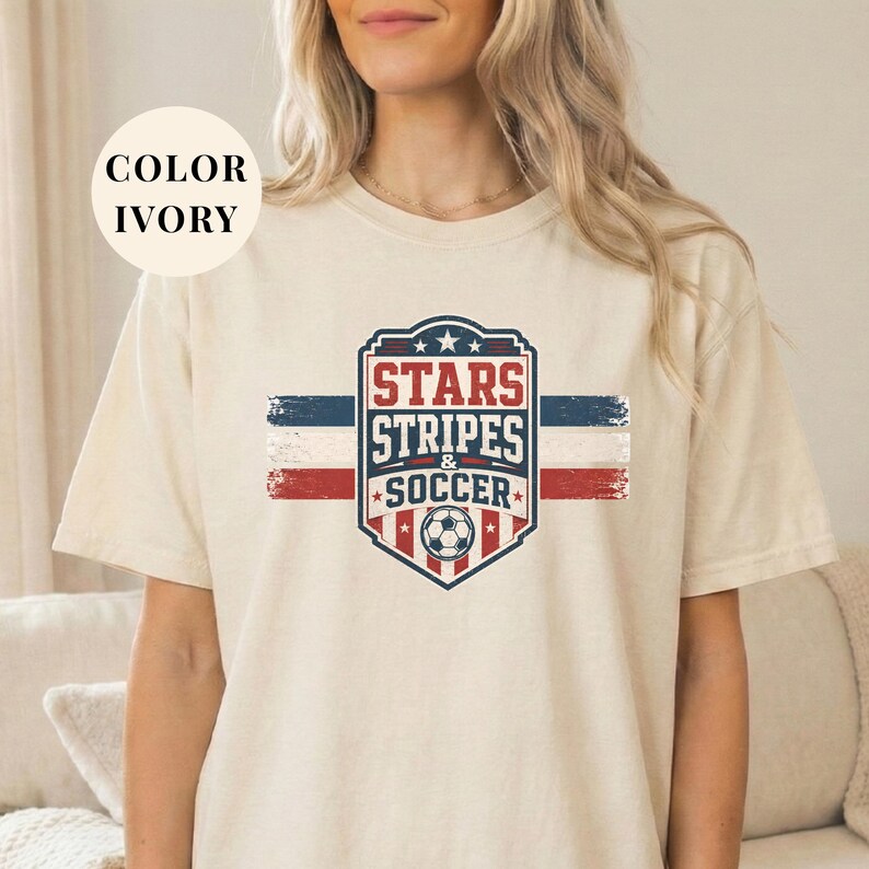 A woman wearing a white t-shirt with a graphic design featuring a shield-shaped logo with the text "STARS STRIPS & SOCCER" and a soccer ball.