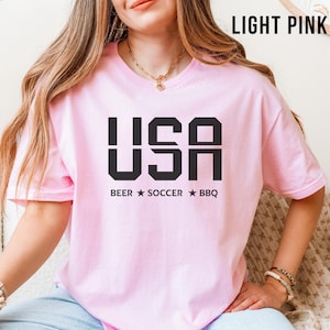 light pink USA T-shirt for Soccer Fans featuring bold block letters and ‘Beer Soccer BBQ’ slogan, patriotic graphic tee for World Cup 2026 and game day wear.