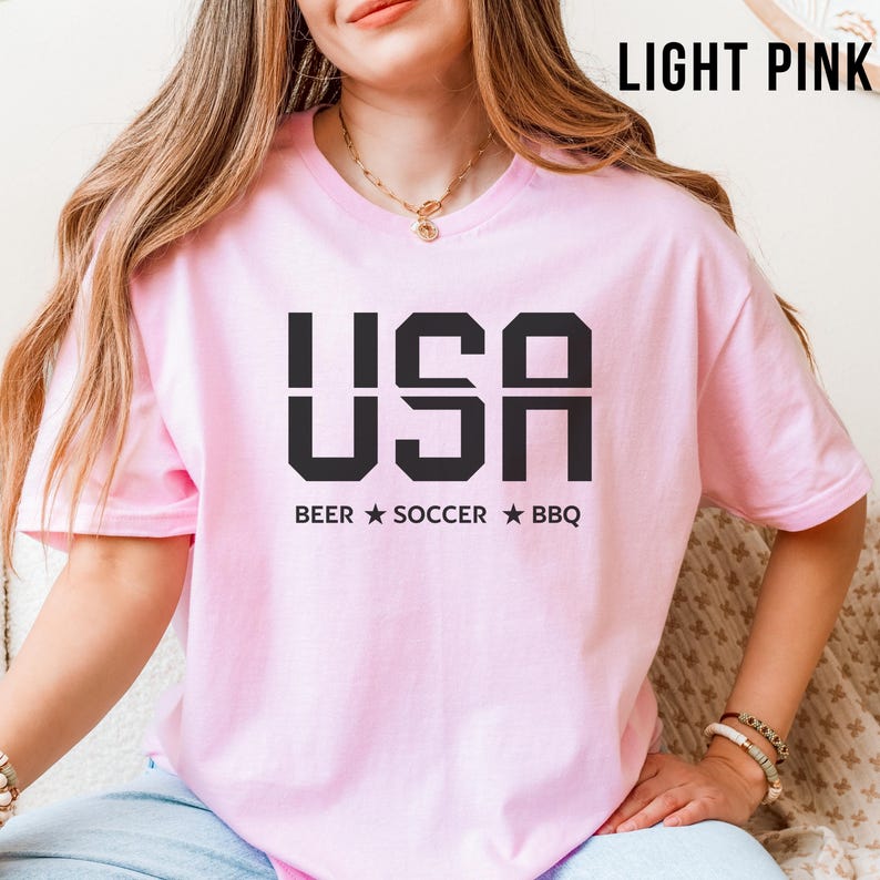 light pink USA T-shirt for Soccer Fans featuring bold block letters and ‘Beer Soccer BBQ’ slogan, patriotic graphic tee for World Cup 2026 and game day wear.