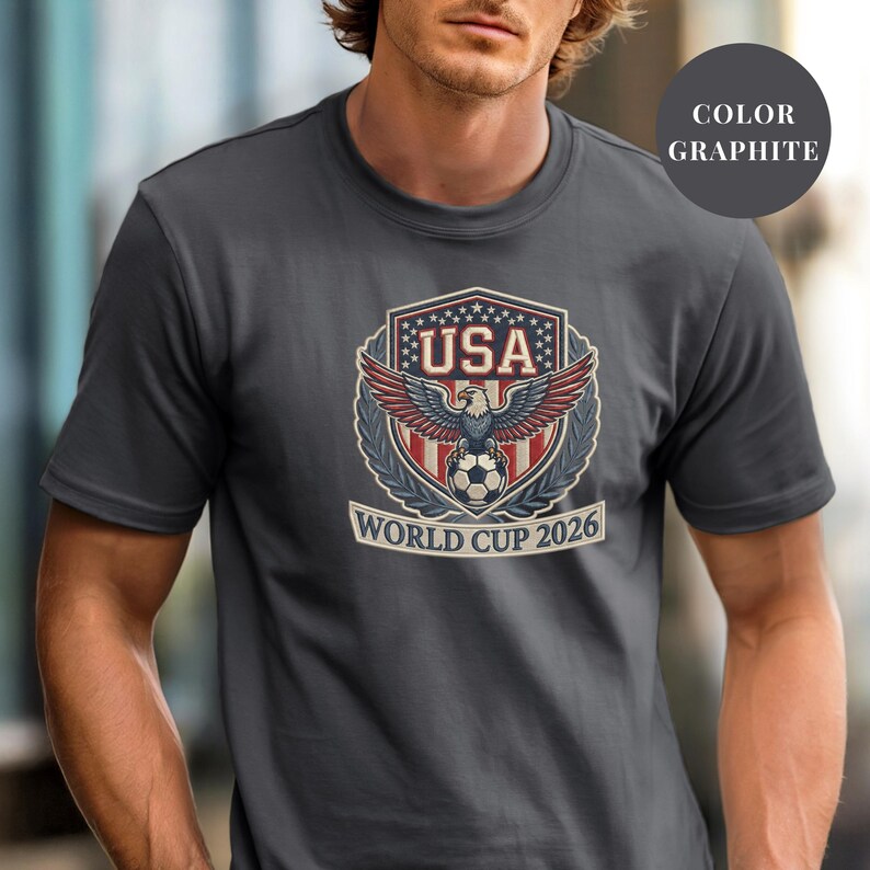 Graphite Comfort Colors 1717 t-shirt featuring a detailed faux embroidery USA World Cup 2026 emblem. The crest shows a bald eagle perched on a soccer ball, surrounded by a laurel wreath and stars. Premium patriotic soccer fan apparel for World Cup 2026.
