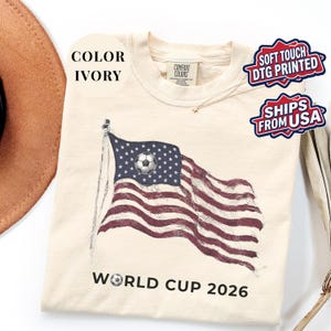 Ivory Comfort Colors 1717 t-shirt featuring a vintage-style waving American flag. The design includes a soccer ball integrated into the stars and "WORLD CUP 2026" bold text. High-quality patriotic sports apparel for United States soccer fans and tournament supporters.