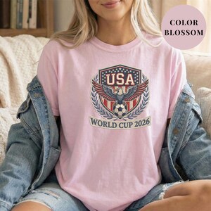 Blossom pink Comfort Colors 1717 t-shirt featuring a detailed faux embroidery USA World Cup 2026 emblem. The crest shows a bald eagle perched on a soccer ball, surrounded by a laurel wreath and stars. Premium patriotic soccer fan apparel for World Cup 2026.