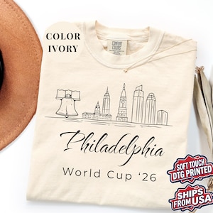May include: Ivory-colored t-shirt featuring a black line drawing of the Philadelphia skyline, including the Liberty Bell. The word "Philadelphia" is written in cursive, with "World Cup '26" below. The shirt is made of Comfort Colors material.