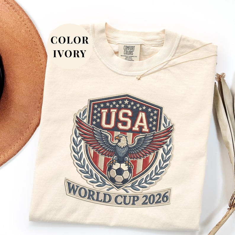 Ivory Comfort Colors 1717 t-shirt featuring a detailed faux embroidery USA World Cup 2026 emblem. The crest shows a bald eagle perched on a soccer ball, surrounded by a laurel wreath and stars. Premium patriotic soccer fan apparel for World Cup 2026.