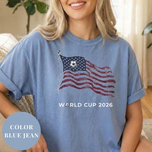 Blue jean Comfort Colors 1717 t-shirt featuring a vintage-style waving American flag. The design includes a soccer ball integrated into the stars and "WORLD CUP 2026" bold text. High-quality patriotic sports apparel for United States soccer fans and tournament supporters.