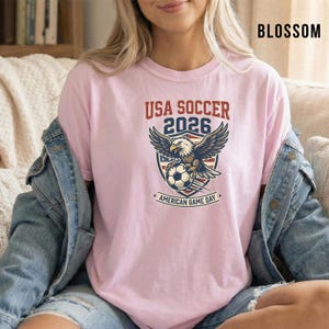 May include: A light pink t-shirt featuring a graphic for USA Soccer 2026. The design includes an eagle, soccer ball, and the text "American Game Day". The word "Blossom" is printed in black in the upper right corner.