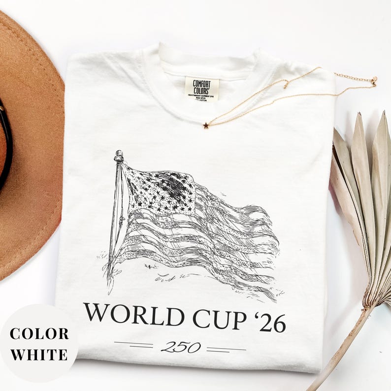 White Comfort Colors 1717 t-shirt featuring a minimalist vintage sketch of a waving American flag. Includes "WORLD CUP '26" and a stylized "250" text celebrating the USA Semiquincentennial. Premium patriotic sports apparel for United States soccer fans and 2026 host nation supporters.