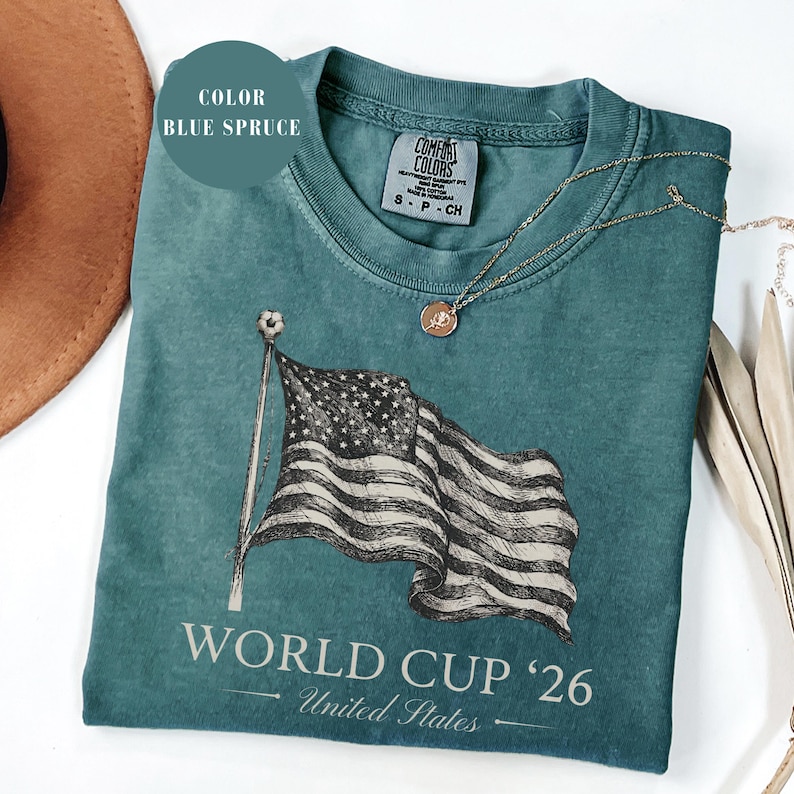 Blue spruce Comfort Colors 1717 t-shirt featuring a vintage hand-drawn sketch of a waving American flag. Includes "WORLD CUP '26" and "United States" text, with a subtle soccer ball detail on the flagpole. High-quality patriotic soccer apparel for the 2026 world tournament fans.