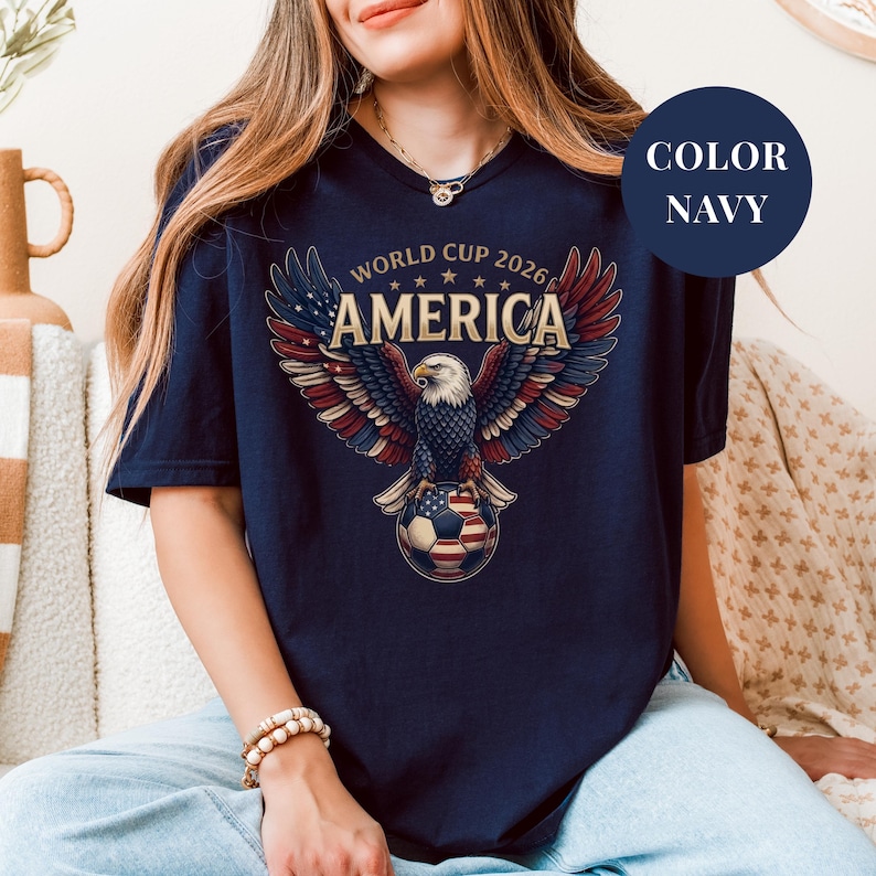 Navy t-shirt featuring a patriotic World Cup 2026 America design. A majestic bald eagle with red, white, and blue wings grasps a USA flag pattern soccer ball below "WORLD CUP 2026 AMERICA" text. Premium sports apparel for United States soccer fans and tournament supporters.