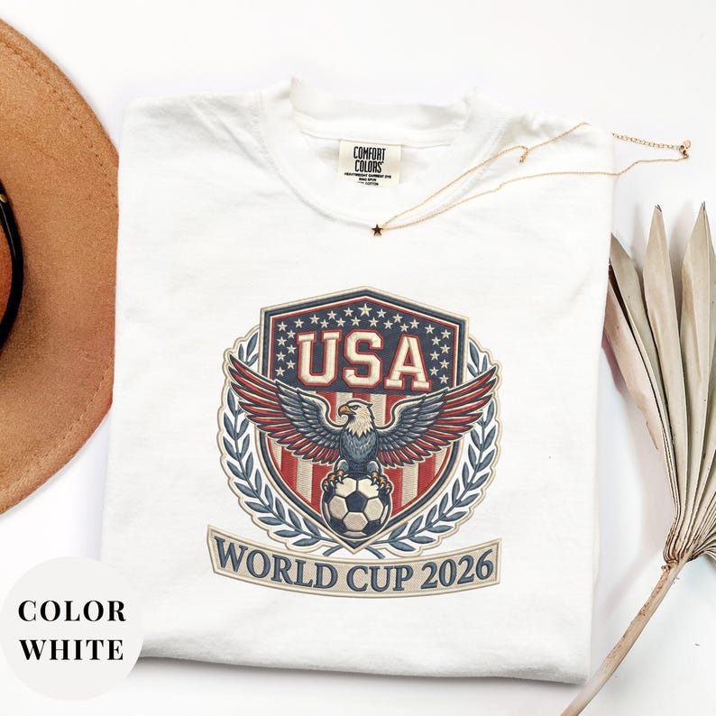 White Comfort Colors 1717 t-shirt featuring a detailed faux embroidery USA World Cup 2026 emblem. The crest shows a bald eagle perched on a soccer ball, surrounded by a laurel wreath and stars. Premium patriotic soccer fan apparel for World Cup 2026.