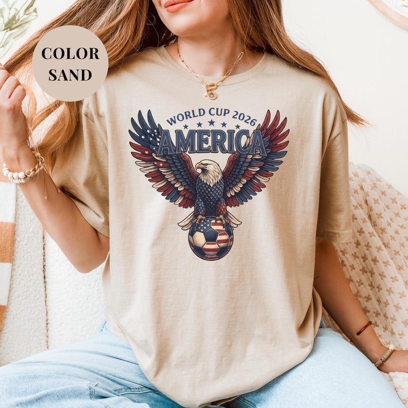 Sand t-shirt featuring a patriotic World Cup 2026 America design. A majestic bald eagle with red, white, and blue wings grasps a USA flag pattern soccer ball below "WORLD CUP 2026 AMERICA" text. Premium sports apparel for United States soccer fans and tournament supporters.
