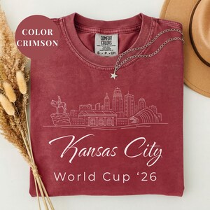 Crimson Comfort Colors T-shirt featuring a minimalist white line art Kansas City skyline design, elegant Kansas City script lettering, and World Cup ’26 text, styled as a soccer travel souvenir shirt for Kansas City fans, football lovers, and 2026 World Cup city apparel.