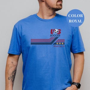 A man wearing a blue t-shirt with a graphic design featuring a soccer ball and the text "US SOCCER World Cup 26".