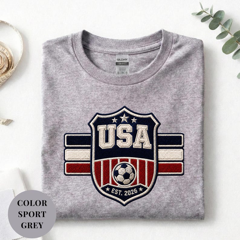 A gray t-shirt with a large embroidered patch that says "USA" and features a soccer ball and stripes, along with the text "EST. 2026" and "COLOR SPORT GREY".