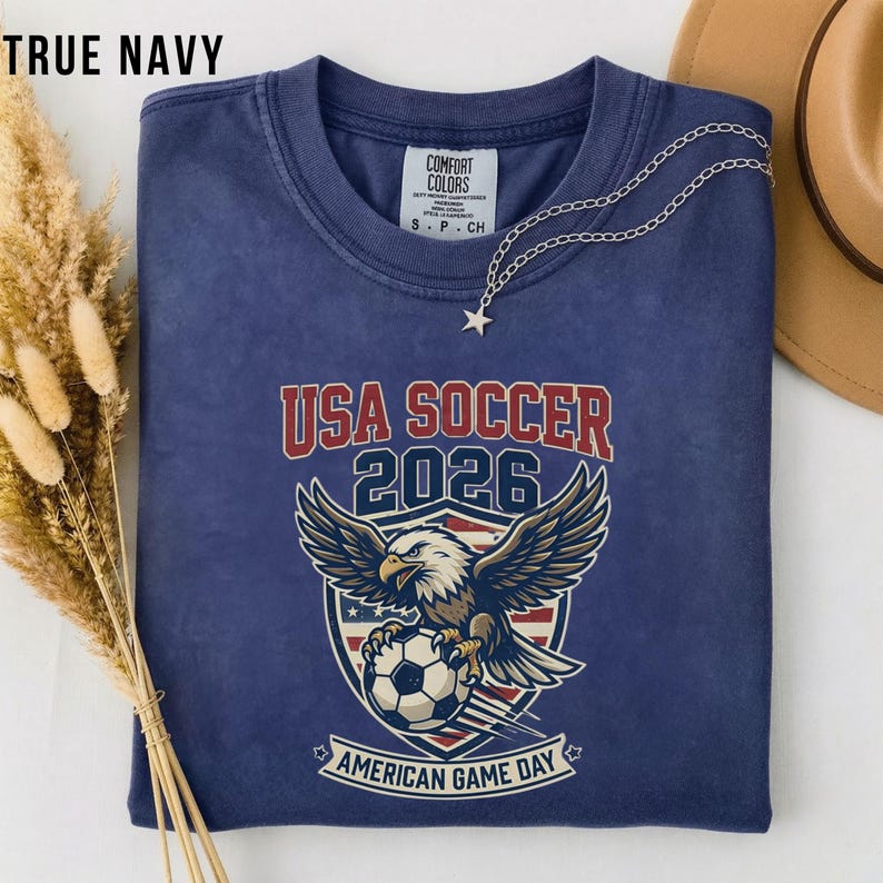 May include: True navy blue t-shirt with "USA SOCCER 2026" text and a graphic of an eagle holding a soccer ball. The shirt also has the text "AMERICAN GAME DAY". A silver necklace with a star pendant is on the shirt.