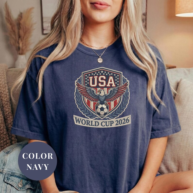 Navy Comfort Colors 1717 t-shirt featuring a detailed faux embroidery USA World Cup 2026 emblem. The crest shows a bald eagle perched on a soccer ball, surrounded by a laurel wreath and stars. Premium patriotic soccer fan apparel for World Cup 2026.