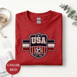 A red t-shirt with a shield-shaped logo that says "USA" and features a soccer ball, along with a label that reads "EST. 2026". The t-shirt is folded and placed on a white surface, with a ruler and some greenery visible in the background.
