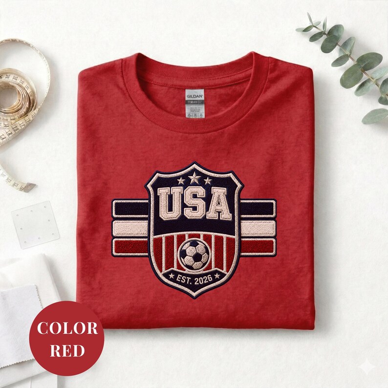 A red t-shirt with a shield-shaped logo that says "USA" and features a soccer ball, along with a label that reads "EST. 2026". The t-shirt is folded and placed on a white surface, with a ruler and some greenery visible in the background.