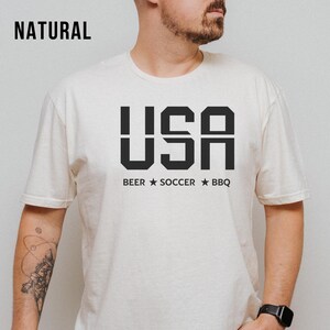 natural USA T-shirt for Soccer Fans featuring bold block letters and ‘Beer Soccer BBQ’ slogan, patriotic graphic tee for World Cup 2026 and game day wear.