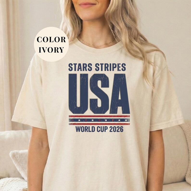 Ivory t-shirt featuring a distressed vintage "STARS STRIPES USA WORLD CUP 2026" graphic. The design includes weathered block lettering for "USA" and a patriotic red, white, and blue banner with stars. High-quality sports apparel for United States soccer fans and world tournament supporters.