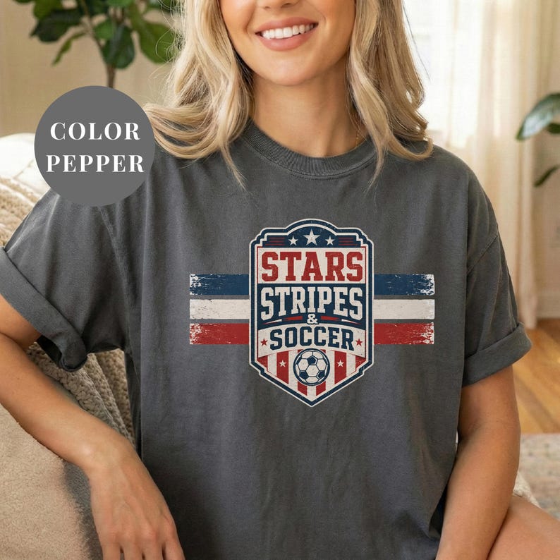 A woman wearing a gray t-shirt with a graphic design featuring a soccer ball and the text "STARS STRIPS & SOCCER" printed on it.