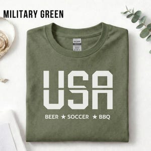 Military Green USA T-shirt for Soccer Fans featuring bold block letters and ‘Beer Soccer BBQ’ slogan, patriotic graphic tee for World Cup 2026 and game day wear.