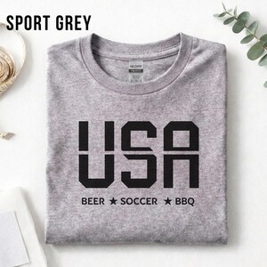 K&ouml;nnte beinhalten: Sportgraues T-Shirt mit dem Wort "USA" in fetten schwarzen Buchstaben. Unter dem Wort "USA" stehen die W&ouml;rter "BEER", "SOCCER" und "BBQ", getrennt durch schwarze Sterne. Das T-Shirt ist ordentlich gefaltet.
