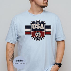 A man wearing a light blue t-shirt with a large shield-shaped logo that says "USA" and features a soccer ball and stripes.