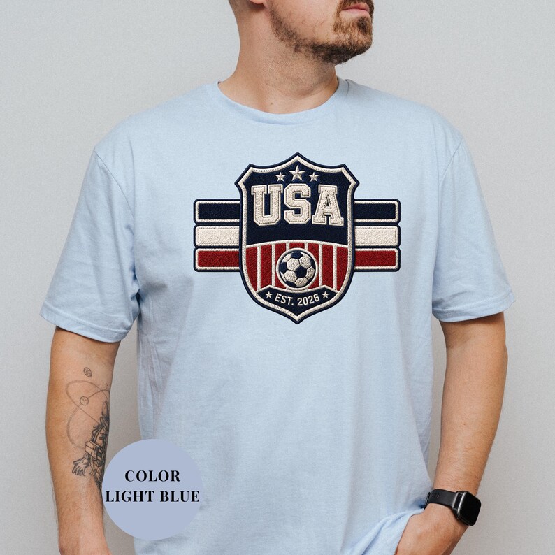 A man wearing a light blue t-shirt with a large shield-shaped logo that says "USA" and features a soccer ball and stripes.