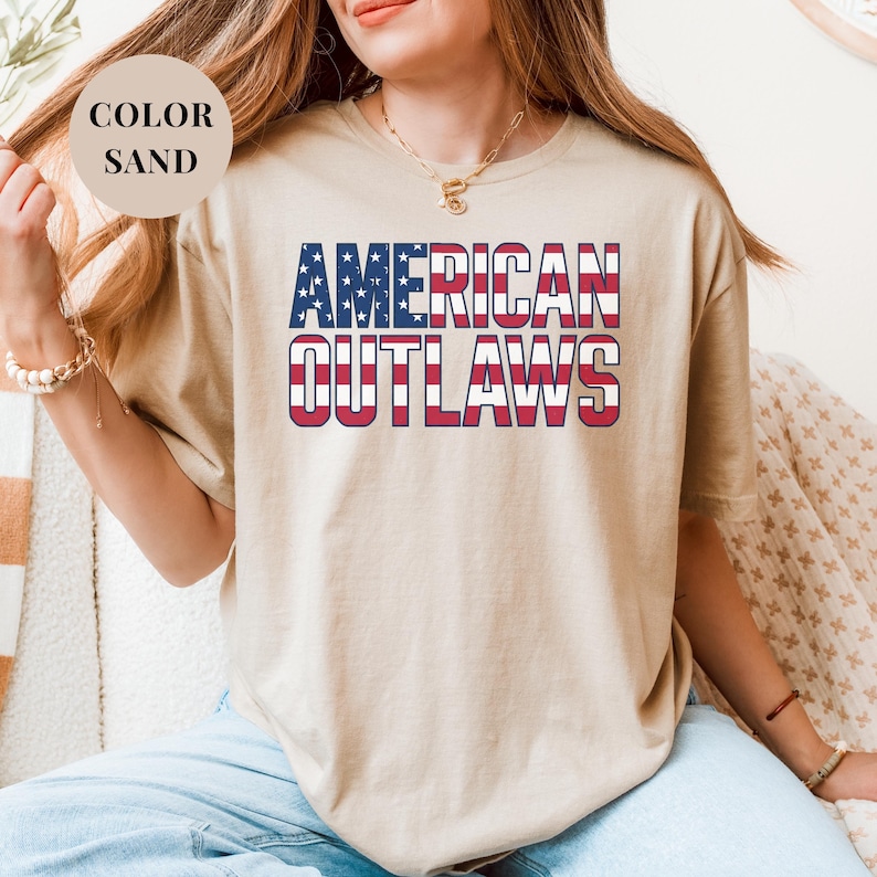 Sand t-shirt featuring a distressed retro design for "AMERICAN OUTLAWS". The bold collegiate block lettering is filled with a patriotic US national flag pattern of stars and stripes. High-quality vintage-style soccer apparel for passionate United States football supporters and tournament fans.