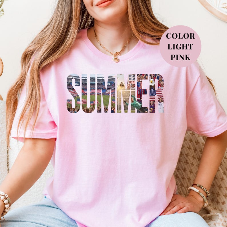 Pink Gildan t-shirt featuring a "SUMMER" photo collage graphic. The bold block letters are filled with USA soccer scenes, including a packed stadium, American flags, cheering fans, and a diving goalie. Patriotic sports apparel for World Cup 2026 supporters.